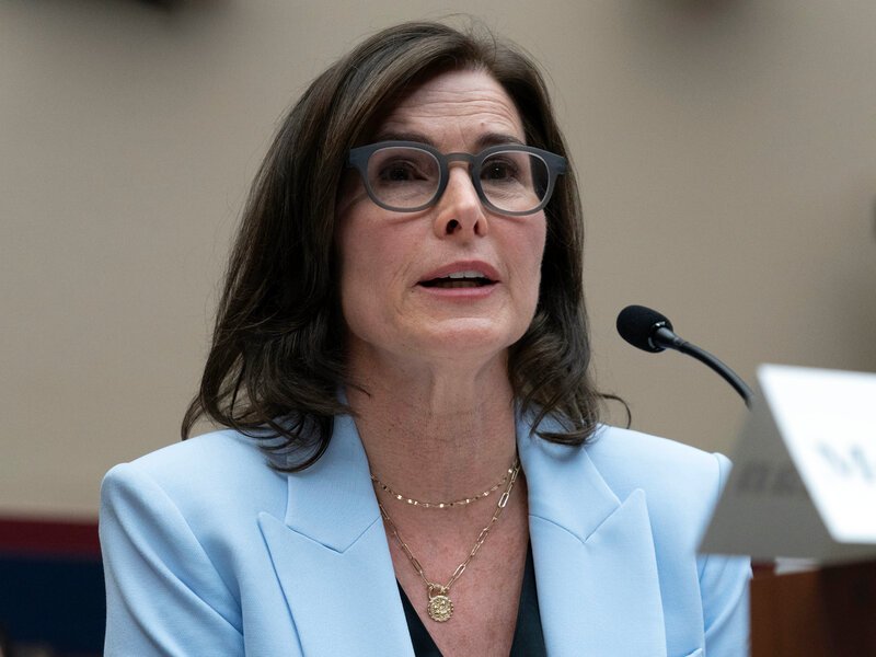 A woman with dark brown hair and glasses, wearing a light blue blazer, speaks into a microphone at a formal event.