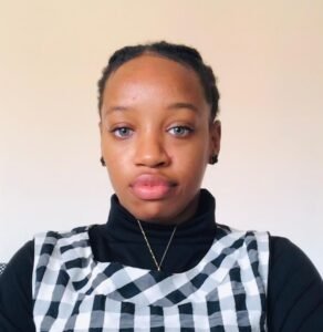 A young woman with braided hair and light eyes wears a black turtleneck under a black and white checkered top, looking at the camera